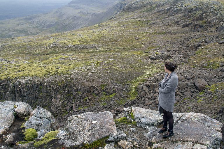 A woman stands, wrapped in a blanket, looking out at the landscape of the Westfjords. Plan the perfect Westfjords itinerary with the help of this guide.