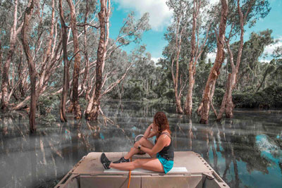 A woman in shorts and a black singlet sits at the end of a boat, looking out onto a creek and gum trees submerged in water in Arnhem Land, one of the best places to visit in Australia.