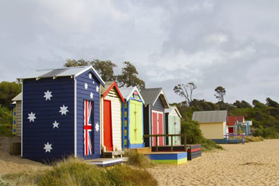 A row of brightly colour beach boxes on a beach in Australia. The first box is painted with the Australian flag. Read through an introduction to Australian slang words.